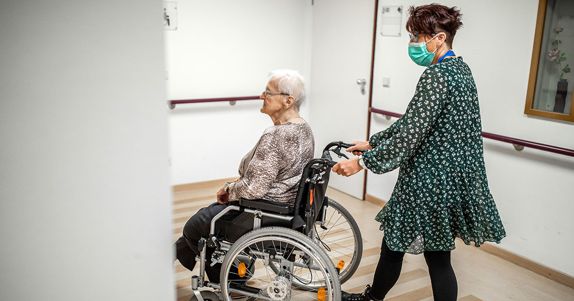 Seniorin im Rollstuhl wird von Pflegekraft durch einen Raum geschoben