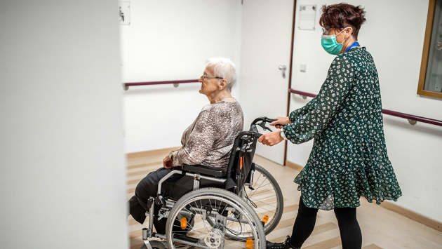 Seniorin im Rollstuhl wird von Pflegekraft durch einen Raum geschoben